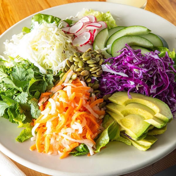 A vibrant salad with shredded cabbage, carrots, red cabbage, avocado, radishes, cilantro, and pumpkin seeds.