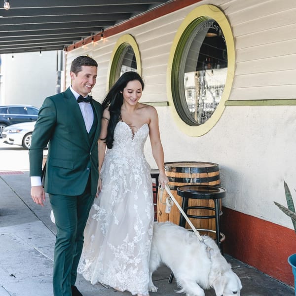 A smiling couple in wedding attire walks a white dog on a leash past a restaurant with round windows.