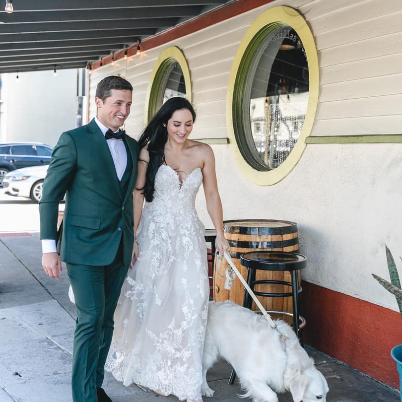 A smiling couple in wedding attire walks a white dog on a leash past a restaurant with round windows.
