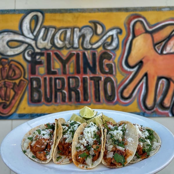 Five tacos with meat, onions, cilantro, and cheese, with lime wedges, in front of a colorful sign.