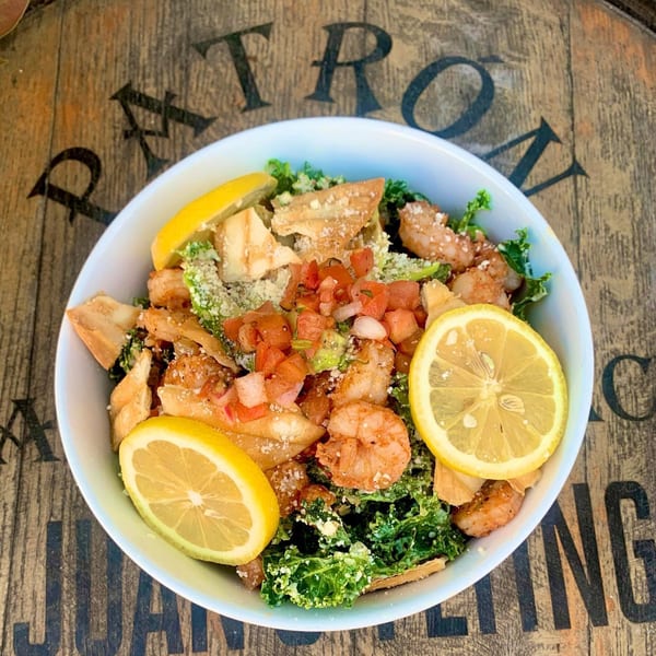 Shrimp and kale salad with pico de gallo, tortilla strips, and lemon slices on a wooden barrel.