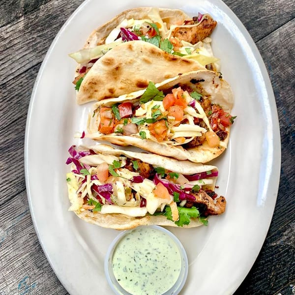 Three tacos filled with chicken, cabbage, and pico de gallo, served with a side of green sauce.