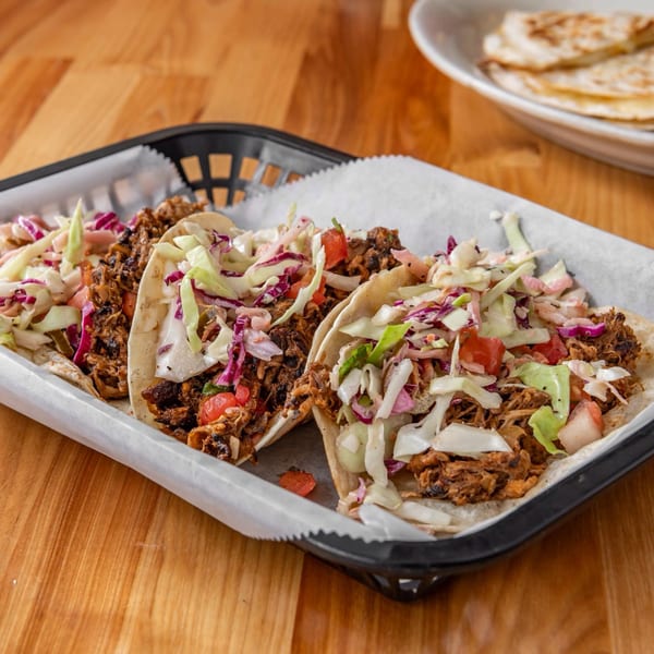Three loaded tacos with shredded meat, cabbage, and tomatoes in a black basket on a wooden table.
