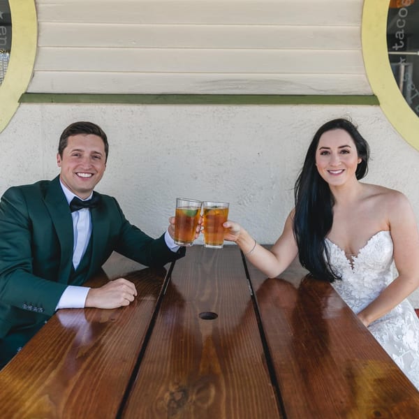 A smiling couple in formal wear toasts with drinks at a wooden picnic table.