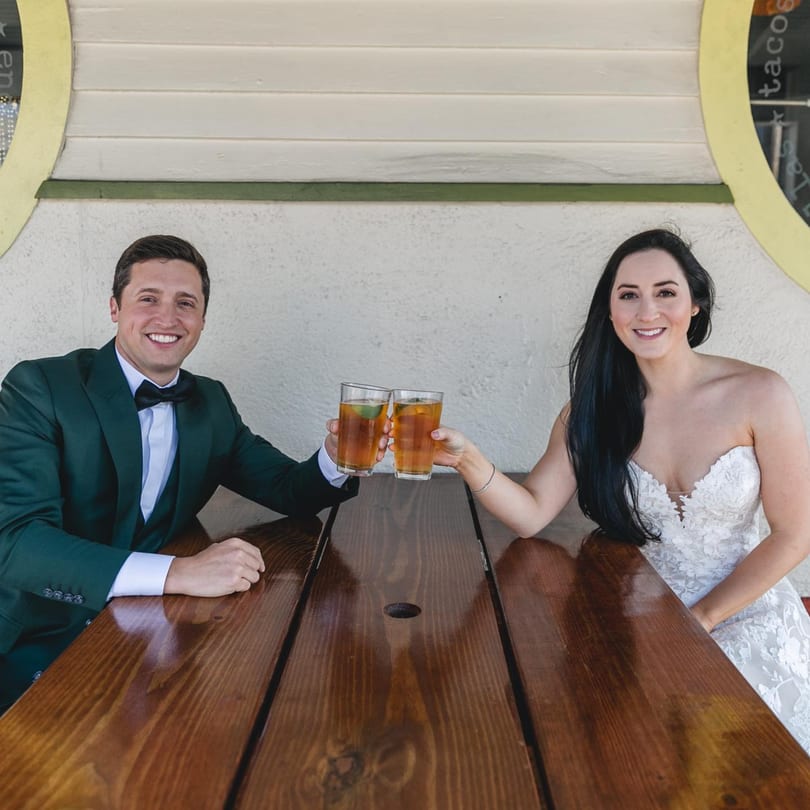 A smiling couple in formal wear toasts with drinks at a wooden picnic table.