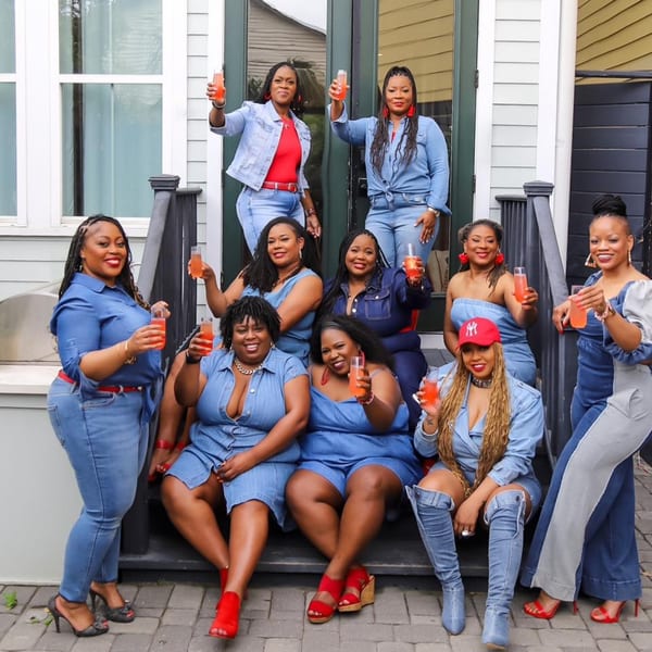Ten women in denim outfits pose on outdoor steps, holding up orange drinks.